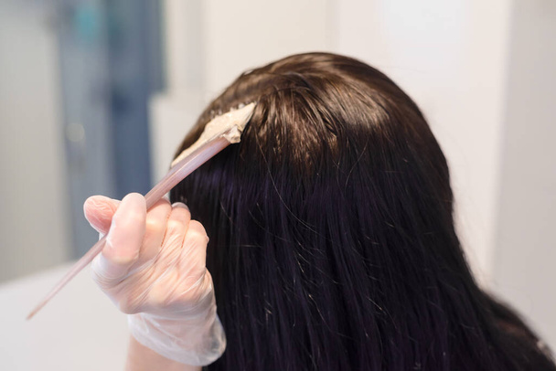 Νεαρή γυναίκα βάφει τα μαλλιά της στο μπάνιο στο σπίτι. Κλείσε. Βαφή μαλλιών με βούρτσα. Χρωματισμός γκρίζων μαλλιών στο σπίτι. Έννοια καραντίνας και πανδημίας. - Φωτογραφία, εικόνα