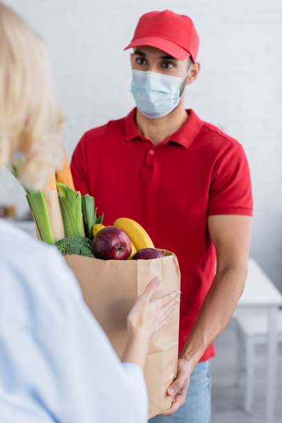 musulmán entrega hombre en máscara médica celebración de alimentos frescos en bolsa de papel cerca de la mujer en primer plano borrosa - Foto, imagen