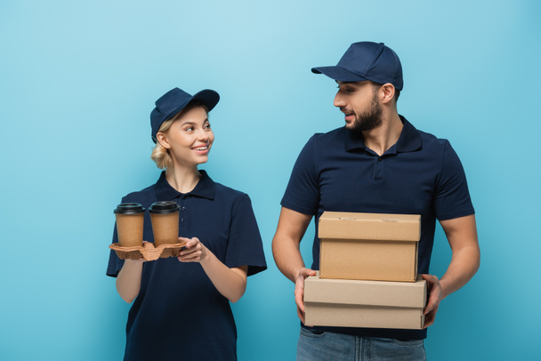 correio multicultural alegre segurando caixas e copos de papel enquanto sorrindo um para o outro isolado em azul - Foto, Imagem