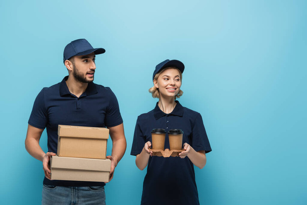 smiling interracial couriers looking away while holding coffee to go and carton boxes isolated on blue - Photo, Image