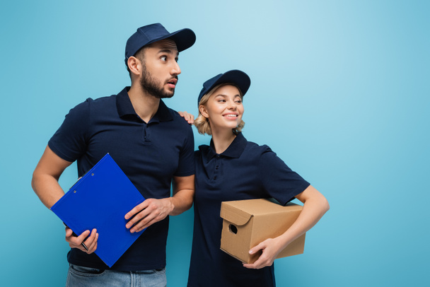 smiling courier holding parcel while looking away together with astonished arabian colleague isolated on blue - Photo, Image