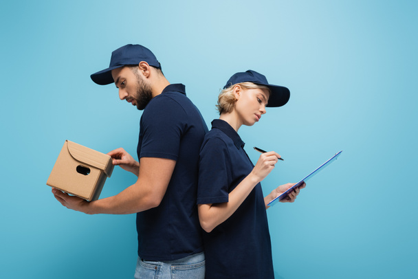 side view of interracial couriers with parcel and clipboard standing back to back isolated on blue - Photo, Image