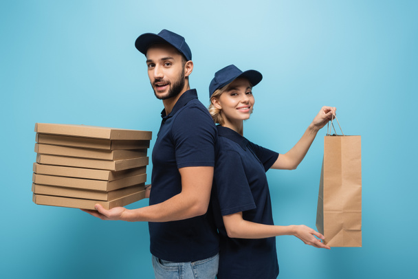 веселые курьеры с коробками для пиццы и бумажным пакетом, смотрящие в камеру на голубом - Фото, изображение