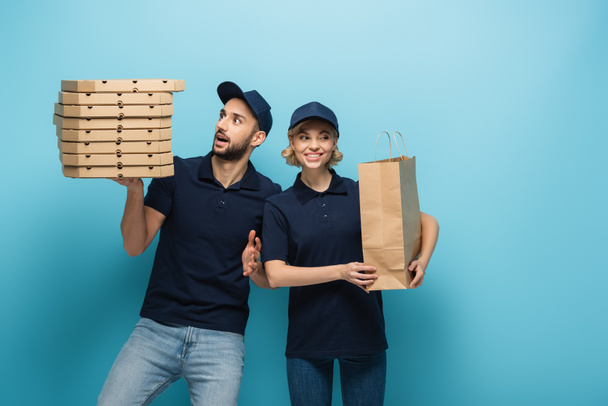 smiling courier with carton packages near excited arabian colleague with pizza boxes on blue - Photo, Image
