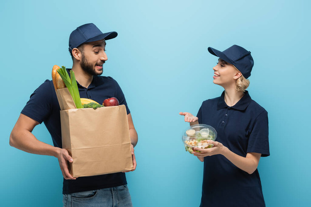 iloinen kuriiri osoittaa lounaslaatikkoon lähellä arabialaista kollegaa, jolla on ruokaa paperipussissa sinisellä - Valokuva, kuva
