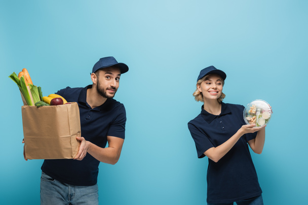 felices mensajeros interracial sosteniendo bolsa de papel y lonchera con comida fresca en azul - Foto, Imagen