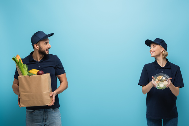 mensajeros multiculturales felices mirándose mientras sostienen paquetes con comida en azul - Foto, imagen