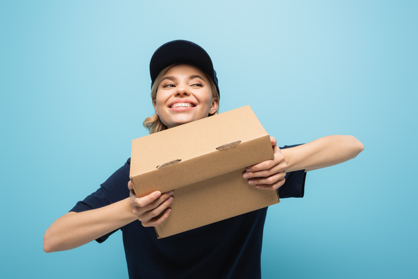 ενθουσιασμένος courier κοιτάζοντας μακριά, ενώ κρατώντας κουτί από χαρτόνι απομονώνονται σε μπλε - Φωτογραφία, εικόνα