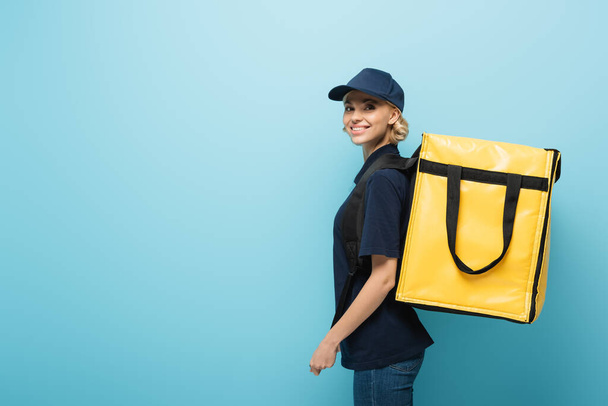 pretty courier with thermo backpack smiling at camera on blue - Photo, Image