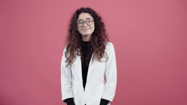 The sincere smile of a mature young woman with curly hair. Young hipster in white jacket and black shirt, with glasses posing isolated on pink background in the studio. - Footage, Video