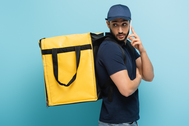 arabian courier with thermo backpack talking on mobile phone on blue - Photo, Image