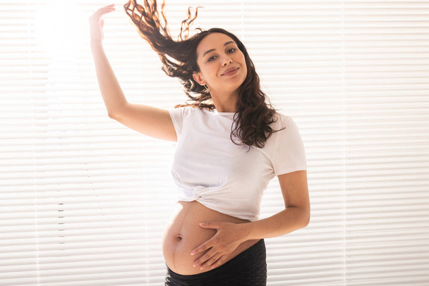 Souriant jeune belle femme enceinte touchant son ventre et ses cheveux et se réjouissant. Concept de santé et de penser à l'avenir en attendant le bébé. Espace de copie - Photo, image