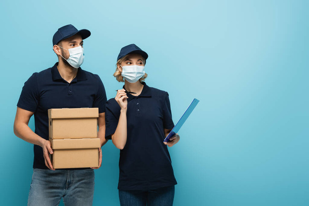 multicultural couriers in medical masks, with parcels and clipboard, looking away on blue - Photo, Image