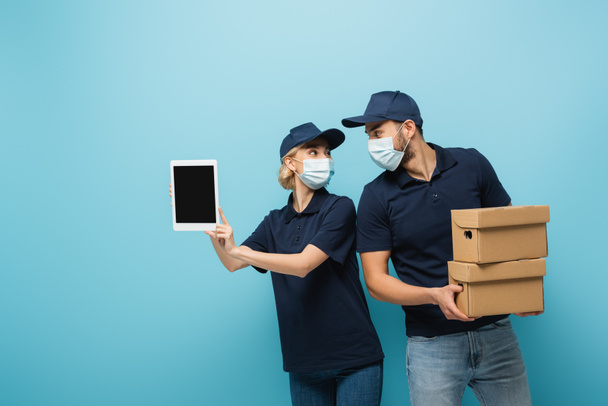 messagers interracial dans des masques médicaux, avec tablette numérique et boîtes en carton, se regardant isolés sur bleu - Photo, image