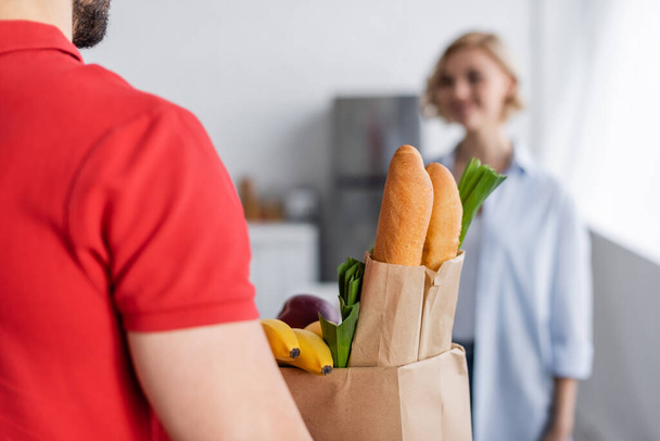 delivery man κρατώντας χάρτινη σακούλα με φρέσκο φαγητό κοντά στη γυναίκα σε θολή φόντο - Φωτογραφία, εικόνα