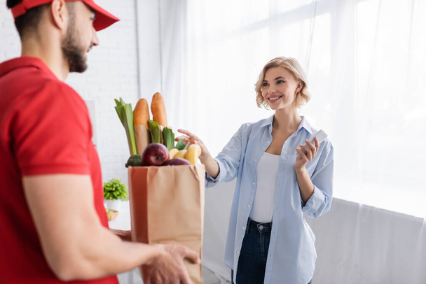 blonde woman taking paper bag with food from arabian courier on blurred foreground - Photo, Image