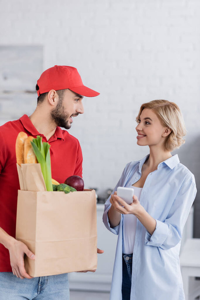 arabian man holding paper bag with fresh food near smiling woman with mobile phone - Photo, Image