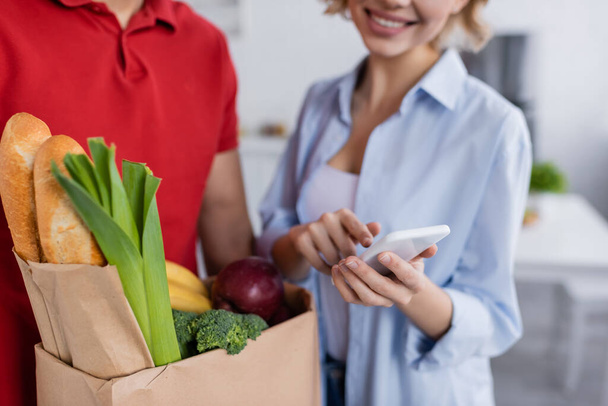 partial view of delivery man with fresh food in paper bag near happy woman pointing at smartphone on blurred background - Photo, Image