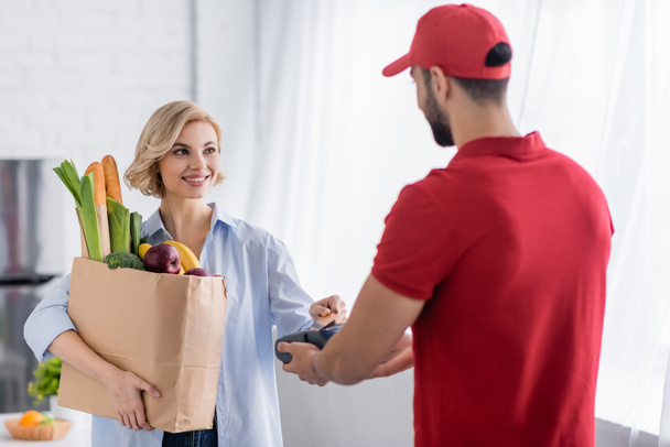 arabian delivery man holding terminal near blonde woman with fresh food in paper bag - Photo, Image