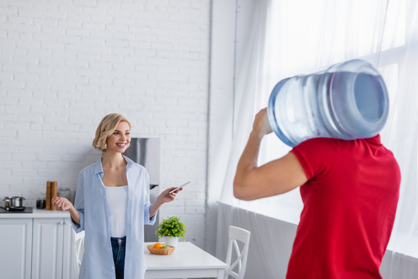 radosna kobieta patrząca na kuriera niosącego dużą butelkę czystej wody - Zdjęcie, obraz
