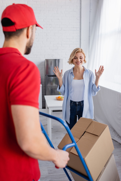 excited woman showing wow gesture near delivery man with boxes on blurred foreground - Photo, Image