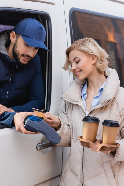 усміхнена жінка з кавою, щоб піти платити замовлення з кредитною карткою біля арабського чоловіка доставки
 - Фото, зображення