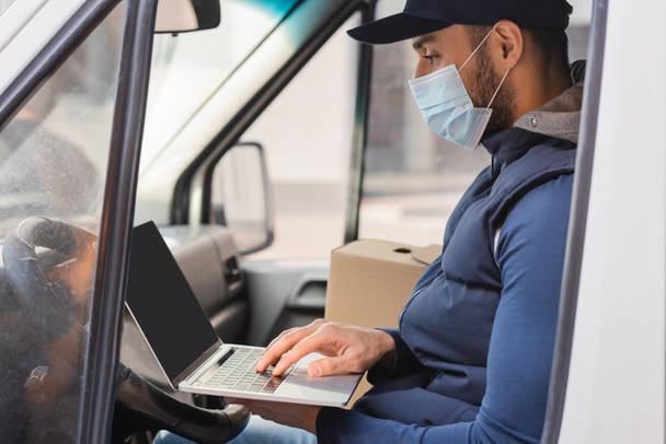 Arabisch levering man in medisch masker typen op laptop tijdens het zitten in de auto - Foto, afbeelding