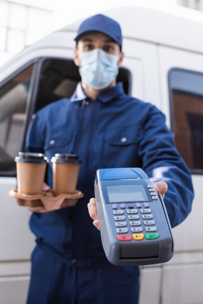 Tıbbi maskeli Arap kurye arka planda bulanık bir ödeme terminaline gitmek için elinde kahve tutuyor. - Fotoğraf, Görsel
