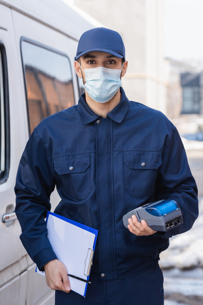 arabischer Kurier in medizinischer Maske mit Zahlungsterminal und Klemmbrett in der Nähe des Autos - Foto, Bild