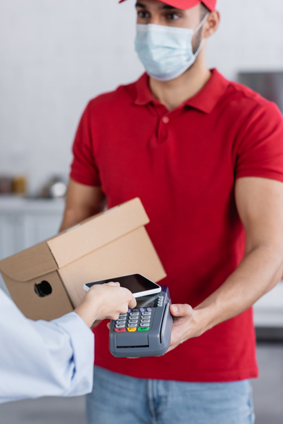 arabian delivery man in medical mask holding payment terminal near woman with smartphone, blurred background - Photo, Image