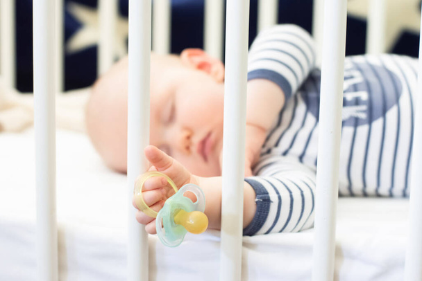 Little baby boy peacefully sleeping in the bed holding pacifier dummy in his hand. Selective focus. - Photo, Image