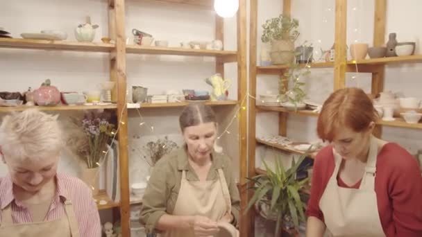 陶器の工房で手作業で土器を作るエプロン姿の二人の先輩の白人女性と若い女性の中出しを傾きながら語り、笑顔で、そして背景にある器のある棚 - 映像、動画