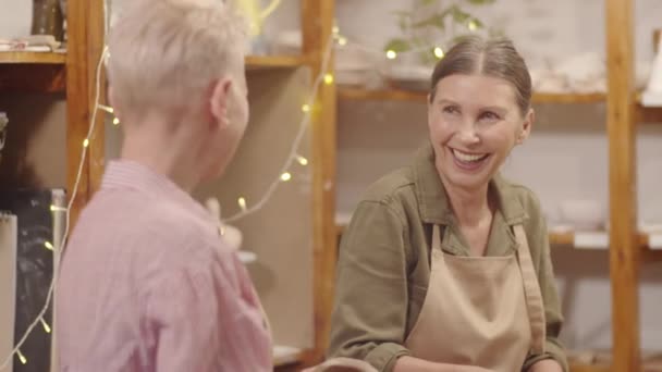 Στιγμιαία φωτογραφία ηλικιωμένης καυκάσιας κυρίας με ποδιά να γελάει και να συνομιλεί με γυναίκα σύντροφο ενώ σμιλεύει πήλινα μπολ σε εργαστήρια κεραμικής - Πλάνα, βίντεο
