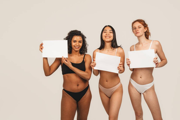 Photo of optimistic cheery young women, multiracial friends wear underwear and hold white plate. Concept natural beauty and girl power - Foto, imagen