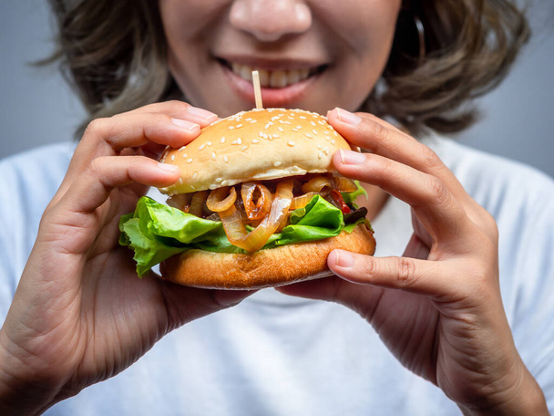 Смачний свіжий саморобний бургер. Одяг красива щаслива азіатська жінка коротке волосся у звичайній білій футболці, що тримає і їсть смачний саморобний гамбургер ізольований на сірому фоні. - Фото, зображення