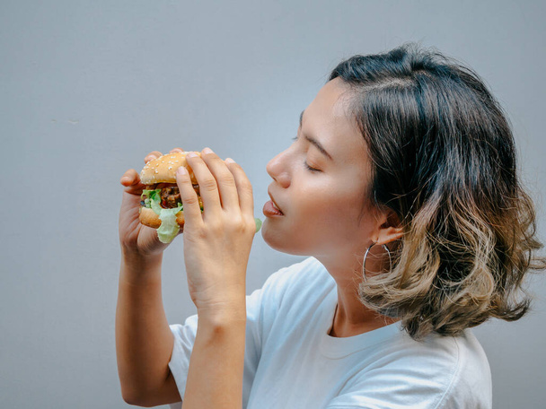 Delicious fresh homemade burger. Beautiful happy Asian woman short hair wearing casual white t-shirt holding and eating tasty homemade hamburger isolated on grey background. - Foto, immagini