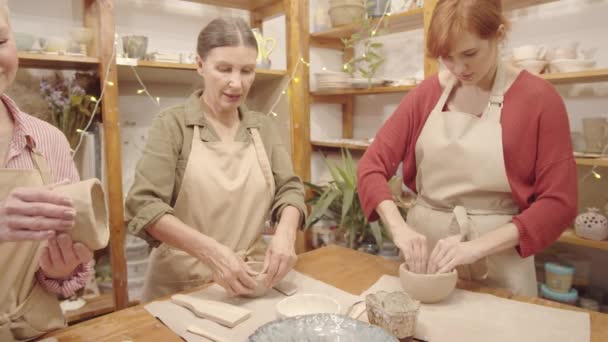 Thigh-up panning shot di giovane insegnante donna caucasica e due studentesse anziane che fanno ciotole di terracotta insieme in studio di ceramica e chatta - Filmati, video