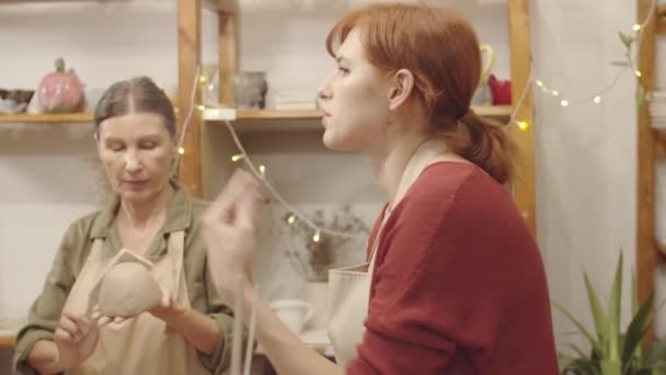 スタジオでのレッスン中に2人の年配の女性と話す若い白人女性陶器教師の胸を打つパンニングショット,そして女性は彼らの粘土ボウルで作業中に聞いて笑顔 - 映像、動画