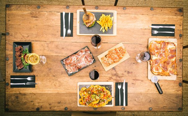 Zenith vista de mesa rústica con nachos, pizza tradicional de jamón, papas fritas y hamburguesas, tataki de atún y un plato de jamón ibérico - Foto, Imagen