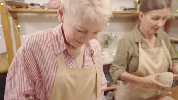Umkippende mittlere Aufnahme einer fröhlichen älteren kaukasischen Dame mit abgeschnittenem grauen Haar, in Schürze und Freundin, die gemeinsam in der Töpferei Tongefäße herstellen - Filmmaterial, Video