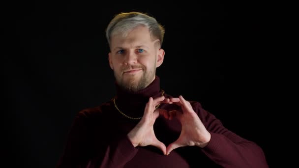 Uśmiechnięty stylowy mężczyzna z niebieskimi oczami ukazujący kształt serca z dłońmi znak kształtu serca, ludzką życzliwość - Materiał filmowy, wideo