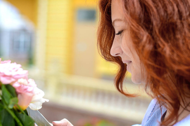 Portret van een roodharig meisje in profiel die lacht, een bericht ontvangt op een smartphone en een boeket bloemen in haar hand houdt - Foto, afbeelding