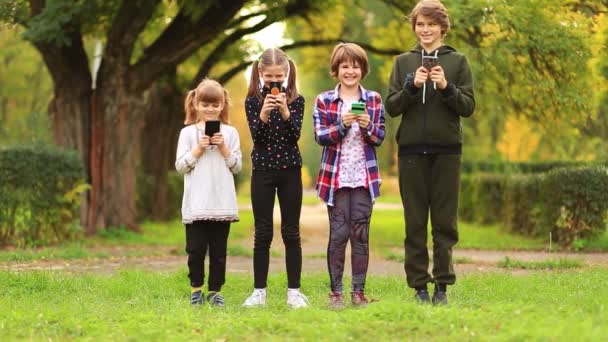 Happy Children ystävä tytöt ryhmä kävellä yhdessä kesällä puistossa ja pelaa internet-peli mobiili älypuhelin. Lapset tekstailevat ulkona kesäpuutarhassa. Bloggaajan elämäntapa - Materiaali, video
