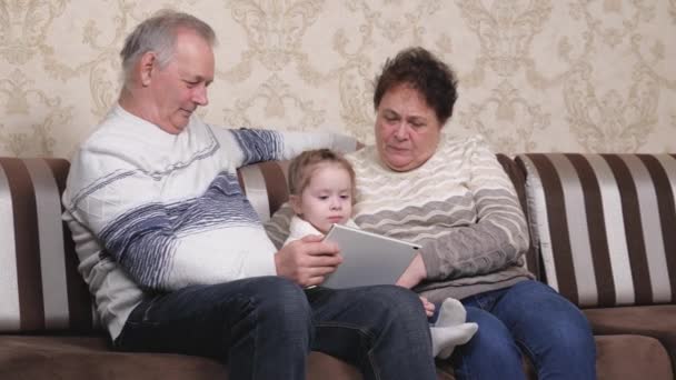 Avô, avó estão brincando com a criança juntos no gadget moderno no quarto. Criança, menina mostra algo em tablet para os avós. Um casal de idosos com neta está usando tablet online - Filmagem, Vídeo
