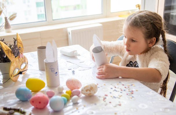 Kız, Paskalya tavşanının kulaklarını yapıştırıyor, Mutlu Paskalyalar için hazırlık yapıyor, çocukların yaratıcılığı, masaya renkli parlak renkli yumurtalar bırakıyor.. - Fotoğraf, Görsel