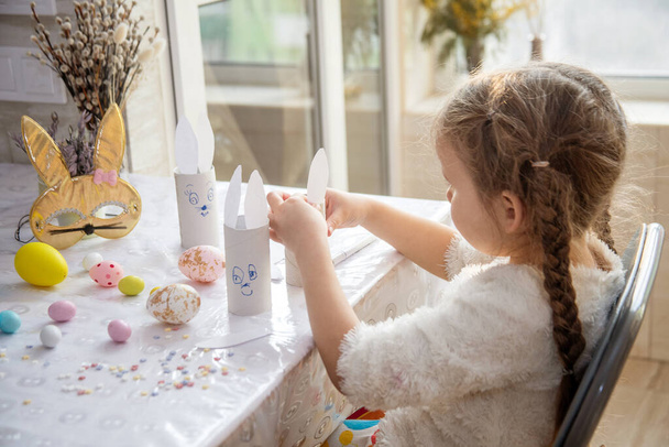The girl glues the ears of the Easter bunny, preparation for Happy Easter, children's creativity, colorful bright painted eggs on the table. - Photo, Image