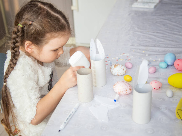 Das Mädchen klebt die Ohren des Osterhasen, Vorbereitung auf Frohe Ostern, Kreativität der Kinder, bunt bemalte Eier auf dem Tisch. - Foto, Bild