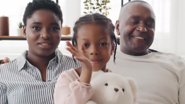 Home ritratto di famiglia, webcam vista tre persone afro americani genitori con figlia guardando la fotocamera con mamma e padre, nero ragazza bambino saluti agitando tenendo giocattolo in mano mostra orsacchiotto a papà - Filmati, video