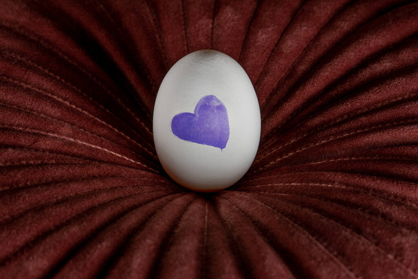 Huevo blanco con corazón azul sobre una almohada roja oscura. Fondo creativo de Pascua - Foto, imagen