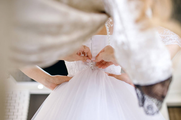 花嫁は結婚式の日に備えてホテルの部屋で美しい白いウェディングドレスを着ています。花嫁介添人は幸せな花嫁がボタンを押すことによって服を着るのを助ける - 写真・画像